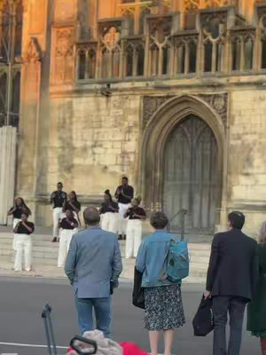 An exterior shot of a large stone building, likely a cathedral or church, with a prominent archway entrance. Several people are standing on the steps, some facing the camera and others towards the arches. A group of people are standing on the stairs in front of the building, they are wearing matching white pants and black shirts, with some holding their hands up in similar gestures. In the foreground, a few individuals are seen walking by and are partially visible. The lighting suggests it could be dusk or early morning.