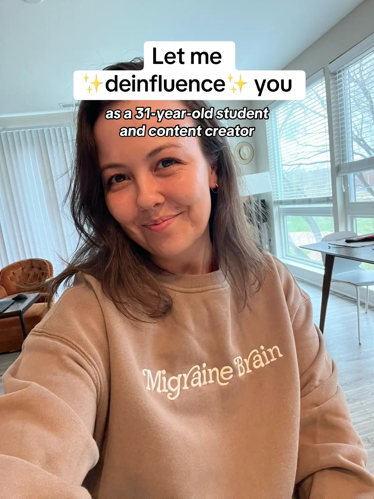 A selfie-style shot of a woman smiling at the camera. She's wearing a tan sweatshirt with the words "Migraine Brain" on it. She has shoulder-length brown hair. Above her, overlaid text says "Let me deinfluence you as a 31-year-old student and content creator."