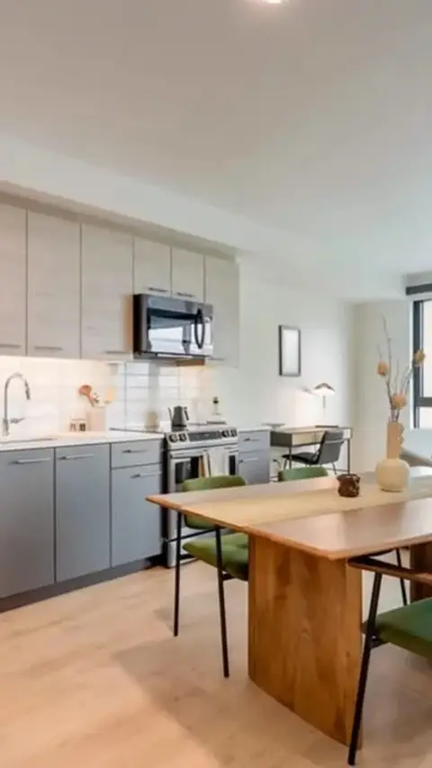 A modern kitchen and dining area. The kitchen has light gray cabinets, stainless steel appliances, and a white tile backsplash. The dining table is wooden with green chairs.