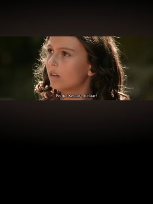 The cover image is a still from a film. A young girl with medium-length brown hair is looking up and to the side with a look of surprise or concern. Her skin is fair. The background is blurred and appears to be outdoors, with tan and green tones suggesting trees or foliage. Text overlayed on the bottom of the image reads: "Pergi | Keluar | Keluar!"