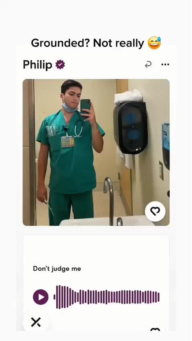 A selfie-style video cover image showing a young man in green scrubs, holding a phone up to take a picture in front of a mirror in what appears to be a hospital setting. The background is white with what looks like a paper towel dispenser on the wall.