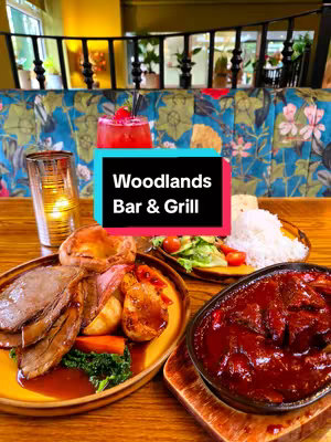 The image shows a table laden with food, including roast beef, Yorkshire pudding, potatoes, and what appears to be a gravy-covered meat dish. A cocktail with a cherry is visible, and a glass with a lit candle. The background is a floral pattern fabric on booth seating, with a glimpse of foliage.