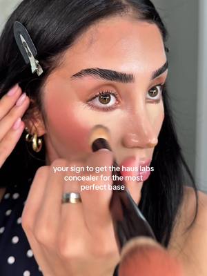 A close-up shot of a person's face, applying makeup. They're holding a makeup brush near their cheek, and the text overlay says, "your sign to get the haus labs concealer for the most perfect base."