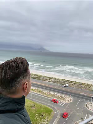 A man from the back looks out onto a coastal scene. His head and shoulders are visible in the foreground with windblown hair. The background shows a beach next to a road with vehicles including some red cars. The sky is overcast.