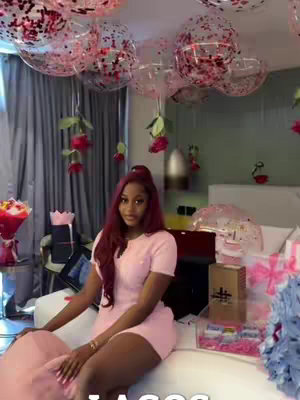 A woman with long red hair in a pink dress sitting on a white couch in a room decorated with pink balloons and roses.