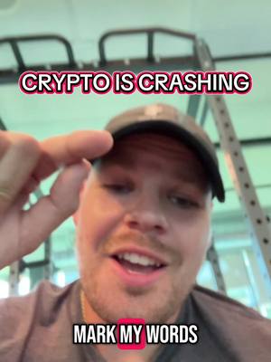 A man in a gray shirt and a black cap is pointing his finger at his head while looking at the camera with a slightly open mouth. The text 'CRYPTO IS CRASHING MARK MY WORDS' is overlaid on the image. The background is blurred but has exercise equipment.