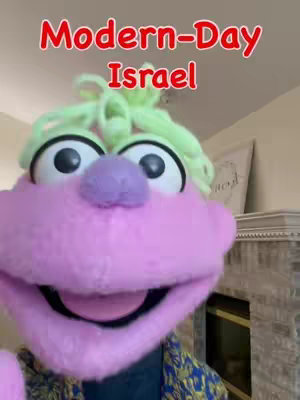 The image features a close-up of a puppet with a large pink face, two big black eyes, a purple nose, and a wide open mouth. The puppet has green fluffy hair-like elements on top. The text overlay at the top reads "Modern-Day Israel" in a red, outlined font. The background includes a partial view of a fireplace and beige wall.