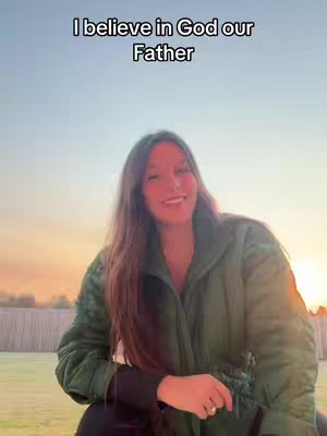 A woman with long brown hair is smiling at the camera. She appears to be outside, with the sky visible behind her, gradually transitioning from blue to orange and yellow, suggesting a sunset or sunrise. She wears a green jacket. Text overlays the image.