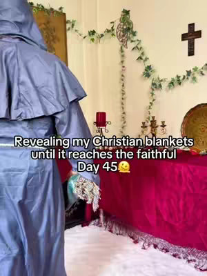 The image shows from the side a person covered in a hooded, blue gown. The person is holding what seems to be a glove made of intricate lace. The background features a wall adorned with greenery, a decorative cross, and a table covered in a vibrant red cloth. Several religious ornaments are present, including a cross and other decorations which give the image a sense of reverence.
