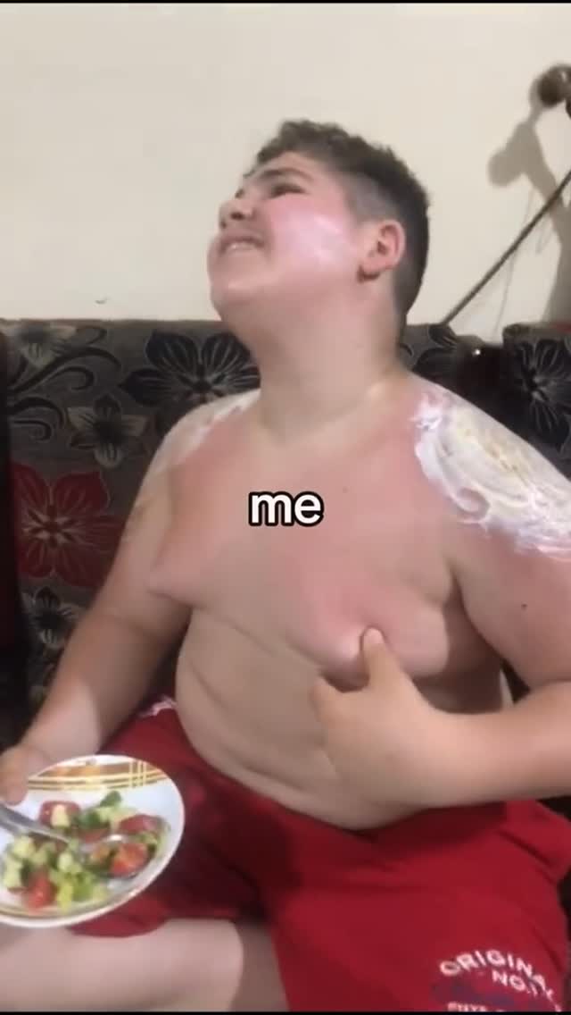 A young person with fair skin is shown from the chest up, seated on a patterned couch. The person's upper body is covered in a white substance, possibly cream or ointment. A plate of salad rests in the person's lap. Red athletic shorts are visible, and a portion of the setting is visible in the background.