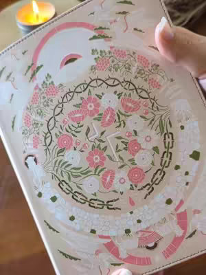 The cover image shows a close-up of a Bible with intricate floral designs and a lit candle in the corner. The Bible is open, and the focus is on the detailed pattern on the cover, which includes pink and white flowers arranged in a circular pattern, surrounded by greenery and decorative elements. A hand is visible, holding the Bible. The background is blurred, suggesting a warm and inviting atmosphere with the presence of the candle.
