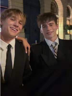 Two young men in suits are standing closely in a candid shot, smiling towards the camera. The person on the left has blond hair and a black tie, and their arm rests on the shoulder of the other person, who has dark hair and a striped tie. The environment suggests a night scene.