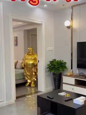 A golden statue of a smiling Buddha stands in a doorway. The room has a modern design with light-colored walls, a black square coffee table, a green plant and a lamp next to a television.