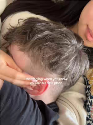 The image shows a close-up of a person's head, partially obscured by another person's arm and hand. The focus is on the side of the head and ear of a person, and a hand is gently resting on their head. The background is blurred, with the person's hair visible. The lighting appears soft, with a warm tone indicating in an intimate setting.