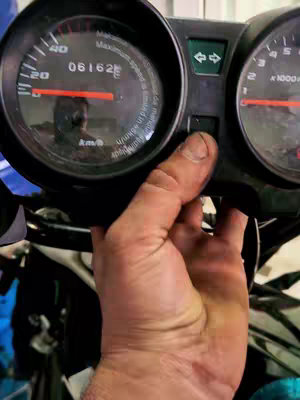 A close-up shot highlights a motorcycle's dashboard. The primary focus is on the speedometer and the visible hand of a person reaching out into the frame. The dashboard features gauges, buttons, and a digital display, with numbers and markings clearly visible. The overall composition emphasizes the mechanical aspect of the motorcycle, with a hand interacting with it.