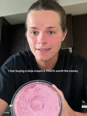 A selfie-style image featuring a woman holding a container of pink, creamy food. She's smiling and looking directly at the camera. The background shows a dark kitchen setting.