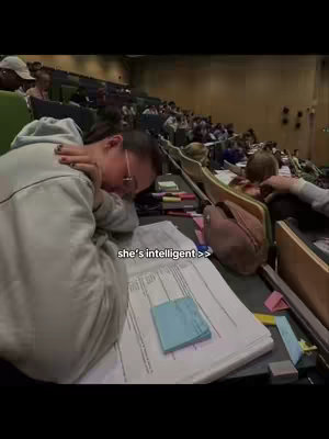 A person resting on a desk in a lecture hall, with documents and a light blue sticky note on the desk. The text overlay reads "she's intelligent >>".