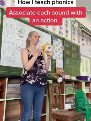 A woman stands in a classroom setting holding up a flashcard with the letter 'W' on it. Text overlays at the top indicate the video is about teaching phonics. The background includes a blackboard with drawings and educational material, suggesting a teaching environment.