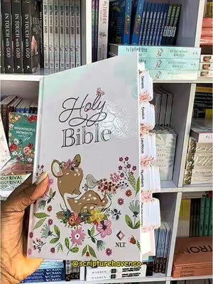 The image presents a close-up of a person's hand holding a white book open to a page with a floral illustration and the words 'Holy Bible' in elegant script. Behind the book are shelves filled with more books and book tabs, creating a sense of a bookstore setting. The overall composition is bright and appealing, focusing on the book's design.
