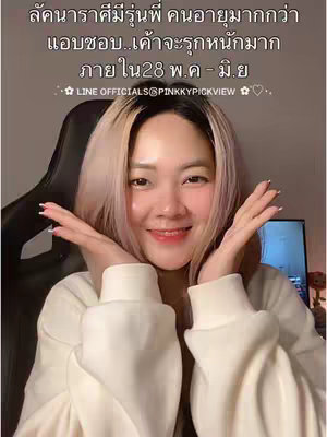 A woman with pale skin and blonde-streaked hair smiles towards the camera, her hands raised near her face in a playful presentation. The background contains a dark office chair, a monitor screen, and indistinct objects. Text overlays provide additional context.