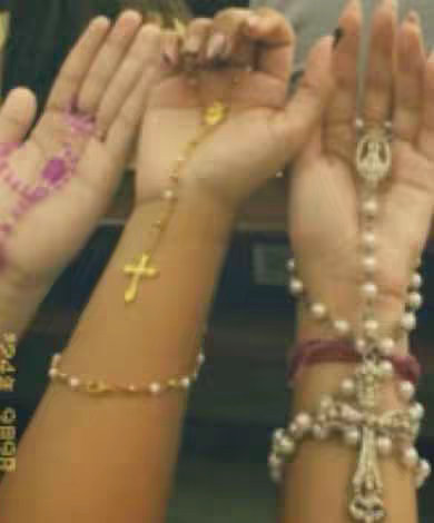 The image shows three arms with hands holding rosaries. The arms have varying skin tones. A gold cross hangs from a rosary, and another wrist has a pearl bracelet. The background is blurred and dark.
