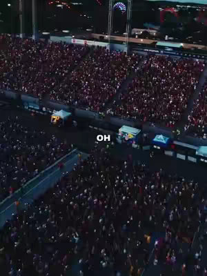 Aerial view of a large outdoor event at night. A massive crowd of people is gathered in a dark area, likely a stadium or open field, illuminated by artificial lights. Structures like a stage or platforms are visible, with a few lit tents. The sky is dark, implying the event is taking place in the evening. The overall composition emphasizes the large scale of the event and the density of the crowd.