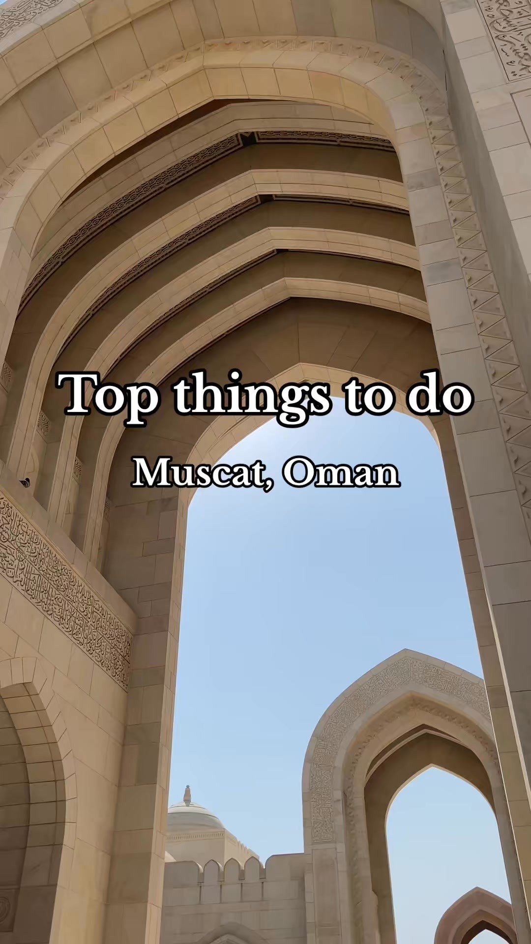 An architectural shot featuring a beige stone archway leading to a bright blue sky. The arch has intricate carvings and is composed of multiple layers. Text overlayed in the middle of the image: Top things to do - Muscat, Oman.