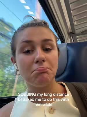A young woman with light brown hair and a neutral expression sits on a train. She wears a white top and has earbuds in her ears. The background shows the interior of a train with windows revealing glimpses of greenery and a bright sky. Text overlaying the image says, "SOBBING my long distance bf asked me to do this with him while."