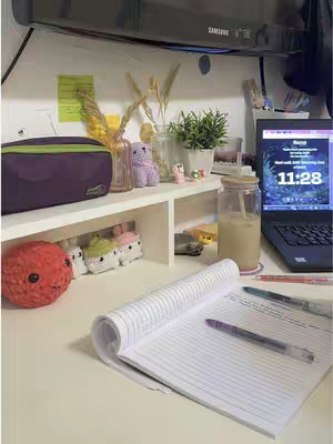 A well-lit desk setup with a laptop, notebook, and various decorative items. A laptop displaying a screen is on the right, a notebook with a pen lies open in the foreground, suggesting a study environment. Various decorative elements such as plants and small figurines are arranged on a shelf. A glass of drink stands near the laptop.
