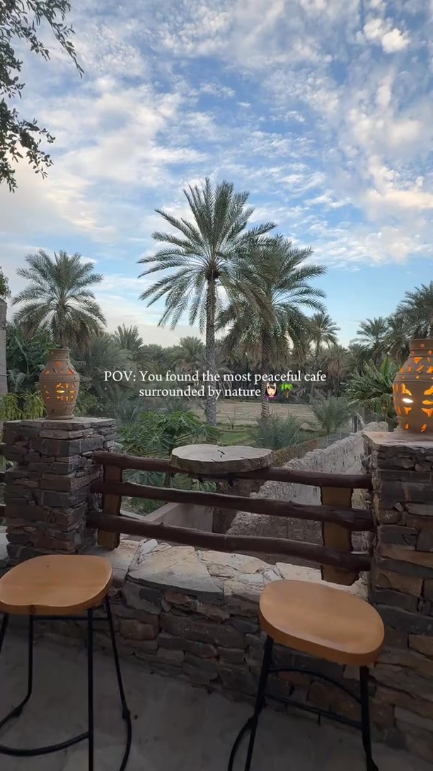 The image features a scenic outdoor view of a cafe setting. A stone and wooden fence with two stools is in the foreground. Beyond the fence lies a landscape with palm trees and other greenery under a cloudy sky. Text overlay on the image states "POV: You found the most peaceful cafe surrounded by nature."