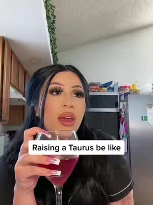 A woman holding a glass of red wine in a kitchen setting. She has dark hair, full makeup, and is looking slightly off-camera. Text overlay in a white box reads, 'Raising a Taurus be like'. The background includes a kitchen with wooden cabinets, appliances, and a glimpse of greenery.