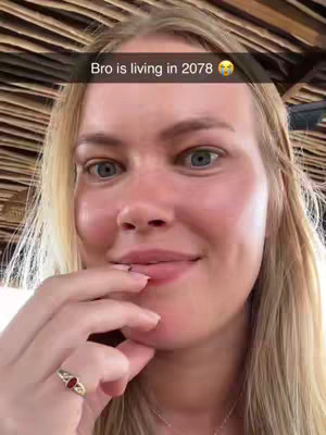 A close-up shot of a woman with blonde hair and fair skin. She looks off-camera with a slightly surprised expression, holding her index finger near her lips. The image has a warm, natural light with a soft focus on her face. A text overlay at the top says, 'Bro is living in 2078'.