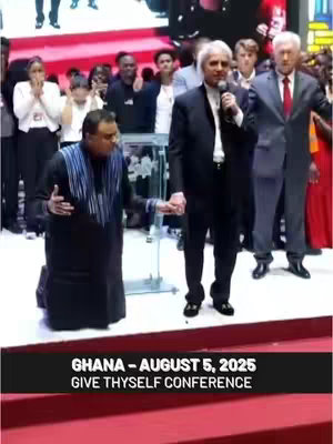The image features a live service with multiple people standing on a stage. The central figures are two men with microphones, dressed in suits, and holding hands. One man is kneeling, appearing to pray. Others are standing behind them, some with their hands raised. The scene is brightly lit with a red carpet in the foreground and a blurred background.