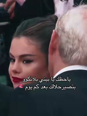 The image shows Selena Gomez, looking over her shoulder with a slight smile, seemingly talking or listening to someone outside the frame. Her lips are a striking red, contrasted against her dark hair. She's in a setting that implies a formal event, with a person partially visible next to her.