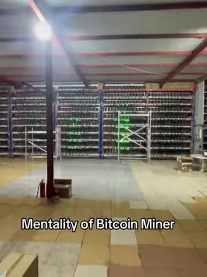 An indoor shot of a large warehouse filled with rows of servers or computers, presumably for Bitcoin mining, is seen from a slightly low angle. The ceiling has exposed metal beams and fluorescent lighting, and the area is well-lit. The foreground is mostly empty with a few cardboard boxes. Text at the bottom reads 'Mentality of Bitcoin Miner'.