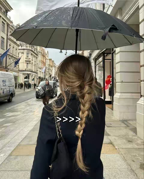 A woman with blonde hair in a braid stands on a city street holding a black umbrella. The background shows buildings and vehicles, and the sky is overcast.