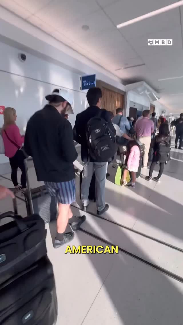 An airport scene with several people, some with luggage, walking through a brightly lit hallway. A person in the foreground is wearing blue and white striped shorts.