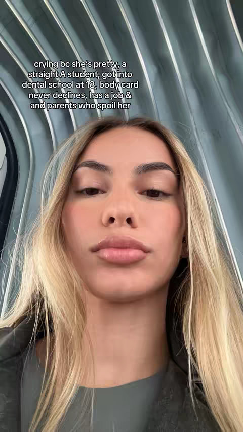 The cover image is a selfie-style video frame featuring a fair-skinned woman with blonde hair. The background is a blurred, metallic, lined surface, possibly part of a building's interior. The woman is looking directly at the camera with a neutral expression; her lips are slightly parted. Text is overlaid at the top, written in a sans-serif white font, and consists of multiple lines of text describing the woman's attributes and situation.