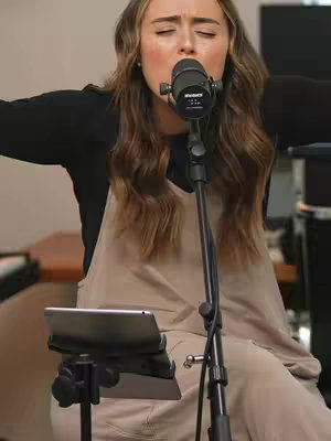 A woman with long brown hair wearing a black top and tan overalls is singing into a microphone. Her eyes are closed, and her arms are outstretched. Behind her is a blurred background of a room with equipment.
