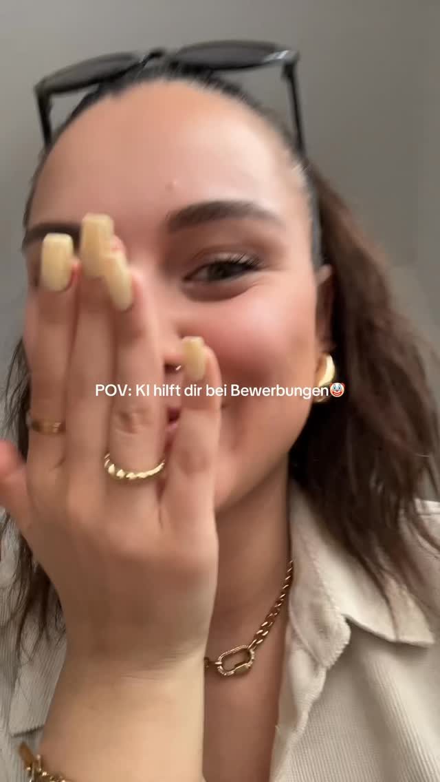 A selfie-style shot with a person, likely a woman, partially covering her face with one hand. She's wearing a light-colored collared shirt and gold jewelry including earrings, a necklace, and rings. The background is indistinct but appears to be an indoor setting. Sunglasses are perched atop her head.