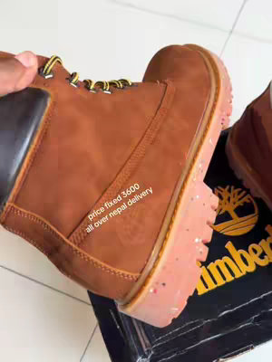 A close-up shot showcasing a brown Timberland boot being held, with text overlayed indicating the price and delivery details. The boot rests over a black and yellow Timberland shoe box.