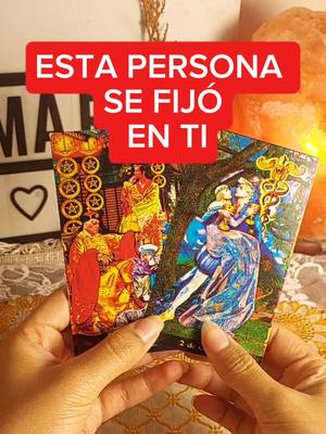 A close-up shot of hands holding tarot cards. A red rectangular text overlay that says "ESTA PERSONA SE FIJO EN TI" (this person noticed you) covers the tarot cards. A light and an object are in the background. The composition is well-lit with a focus on the hands and cards.