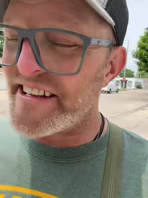 A medium shot of a man's face from the nose up, with the upper half of his body visible. He wears glasses and a baseball cap. His expression is intense, with lips slightly parted. He is in an outdoor setting with a blurred background.