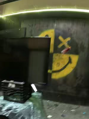 The image captures a moment after impact. A shattered glass surface is in the foreground, with fragments scattered. A bottle appears to be suspended in the air. Behind this, a wall features a painted smiley face. The composition is slightly blurred, giving a sense of action.