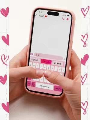 A person's hands holding a pink iPhone displaying a custom keyboard interface for typing on the screen. The keyboard's keys are pink, as is the phone casing. Pink hearts are scattered in the background.