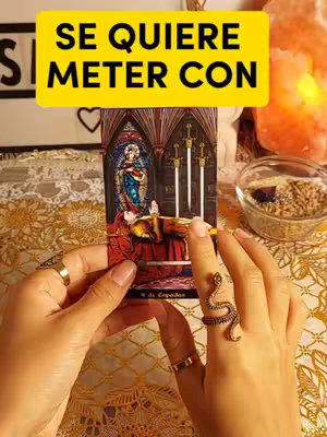 The cover image shows a person's hands holding a tarot card. The card depicts three swords piercing a person lying down in a gothic setting. The person is wearing rings and accessories. A yellow banner with white text is placed on the top. The background includes a softly lit, orange-colored object. The table is covered with an ornate golden cloth. There is a bowl with seeds on the right next to a lamp.