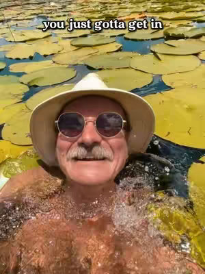 A selfie-style image featuring a man in a white hat and sunglasses, positioned in a pond filled with lily pads. He's smiling with his upper body submerged in water. The background is filled with circular yellow lily pads.