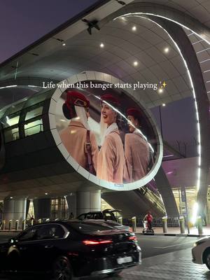 An outdoor shot at dusk or night, featuring a modern architectural structure with a large, circular display showing three women in Emirates airline uniforms. Cars and people are visible in the foreground, with the overall lighting suggesting a city environment.