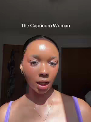 A close-up shot of a woman with smooth, glowing skin. The woman has styled dark hair pulled back, and is wearing a purple top. She has a subtle makeup look with blush on her cheeks and is looking slightly off-camera with a slight smile, as the upper right corner of her mouth is slightly upturned. The text overlay reads 'The Capricorn Woman'. The backdrop is blurred, the light coming in from a window on the left.