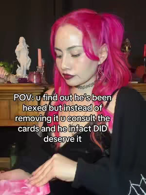 A woman with bright pink hair is centered in the frame, looking slightly off to the side. The text overlay reads, "POV: u find out he's been hexed but instead of removing it u consult the cards and he infact DID deserve it." She is wearing a black top and has dark lipstick. The background is somewhat blurred, with hints of candles and other objects.